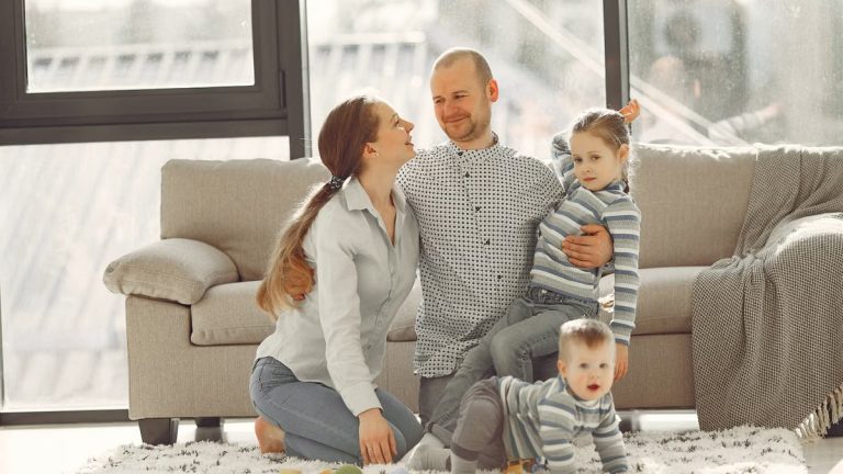 Cheerful mother and father smiling at each other while little cute boy crawling on floor and preschool girl in striped turtleneck raising arm and looking at camera